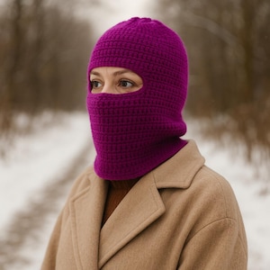 May include: A vibrant magenta balaclava, crafted from a textured knit fabric, covers the head and neck. The balaclava has an opening for the eyes and is worn with a tan coat. The background is a blurred winter scene.