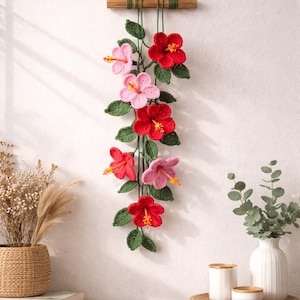 Peut inclure: Décoration murale artisanale avec des fleurs d'hibiscus crochetées en rouge et rose, avec des feuilles vertes. L'arrangement floral est suspendu à une barre en bois avec de la ficelle verte, ajoutant une touche de décoration inspirée de la nature.