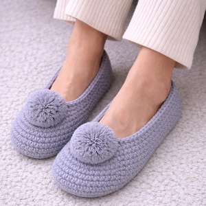 May include: A pair of light purple crocheted slippers with a large pom-pom on each toe. The slippers are on a light-colored carpet. The slippers are a comfortable, cozy style.