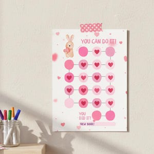 May include: A pink and white chart with a bunny holding a heart, and the text "YOU CAN DO IT!" and "YOU DID IT! NEW BOOK!". The chart has pink circles and heart shapes, taped to a wall.