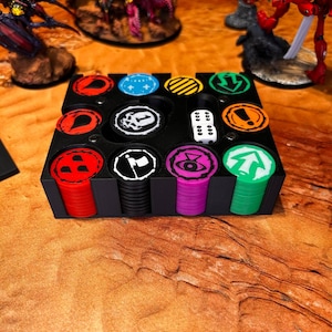 May include: Black plastic game token organizer with various colored tokens and a white die. The tokens are red, blue, yellow, black, purple, and green, each with unique symbols. The organizer is on a wooden surface.