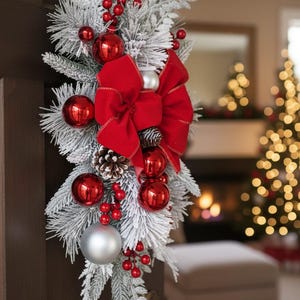 May include: A Christmas door decoration with frosted evergreen branches, red baubles, and a large red velvet bow. Silver and red berries and a pinecone add to the festive look. The decoration is attached to a dark brown door.