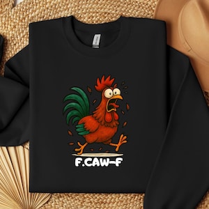 May include: Black crewneck sweatshirt featuring a cartoon rooster with red, orange, and green details, running with an open mouth. The text "F.CAW-F" is printed below the rooster in white. The sweatshirt is on a woven surface.