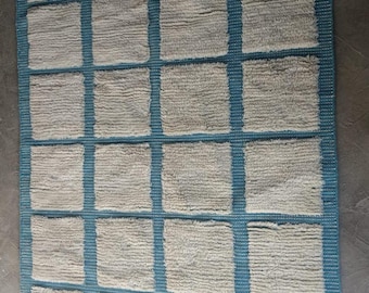 May include: Rectangular rug with a grid pattern. The rug features a light grey, textured surface with a teal blue grid design. The edges are finished with a teal border and fringe.