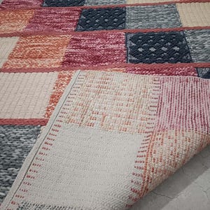 Op de afbeelding: Een kleurrijk patchwork tapijt met een verscheidenheid aan texturen en patronen. Het tapijt heeft vierkanten in roze, oranje, grijs en crème kleuren. De onderkant van het tapijt is zichtbaar en toont een lichter, patroon.