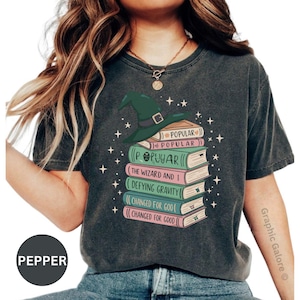 May include: A charcoal gray t-shirt featuring a graphic of a stack of books topped with a green witch hat. The book spines are labeled with the text Popular, The Wizard and I, Defying Gravity, and Changed for Good, surrounded by small white stars.
