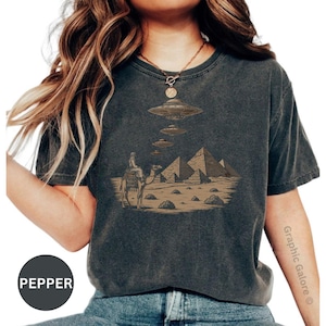 May include: Dark gray t-shirt featuring a sepia-toned graphic of pyramids, a camel rider, and UFOs. The design evokes a sense of mystery and ancient history. The shirt has a classic crew neck and short sleeves.