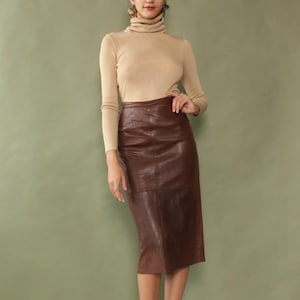 May include: A woman wearing a tan turtleneck sweater and a brown leather pencil skirt. The skirt is knee-length and has a textured appearance. The woman is also wearing strappy sandals.
