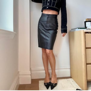 May include: A black leather pencil skirt with a high waist. The skirt is knee-length and has a classic, tailored design. The model is wearing black pointed-toe heels with silver embellishments. The outfit is completed with a black cropped jacket.