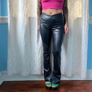 Puede incluir: Pantalones negros de cuero sintético con pernera acampanada. Los pantalones tienen una cintura alta y un acabado liso y brillante. Los pantalones se combinan con sandalias de plataforma verdes.