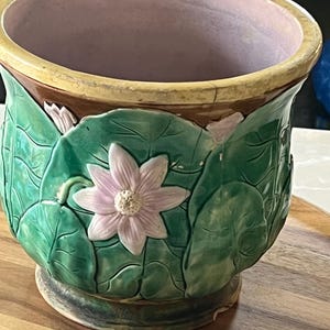 May include: A ceramic planter with a lotus flower design. The planter has a light pink interior, a green exterior with embossed leaves, and a light pink flower. The rim is brown and yellow.
