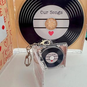 May include: A clear CD case featuring a black and white vinyl record design with the words "Our Songs". A miniature keychain version of the CD is also present. A red heart is on the record label.