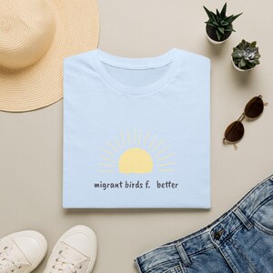 Könnte beinhalten: Ein hellblaues T-Shirt mit einer gelben Grafik einer aufgehenden Sonne und dem in Schwarz gedruckten Text "migrant birds f. better". Das T-Shirt wird zusammen mit einem Strohhut, einer Sonnenbrille, zwei kleinen Sukkulenten und weißen Turnschuhen gezeigt.