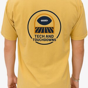 K&ouml;nnte beinhalten: Senfgelbes T-Shirt mit einem marineblauen und orangefarbenen Football-Motiv auf dem R&uuml;cken. Das Design zeigt einen Football, Torpfosten und den Text "TECH AND TOUCHDOWNS". Das T-Shirt hat kurze &Auml;rmel.
