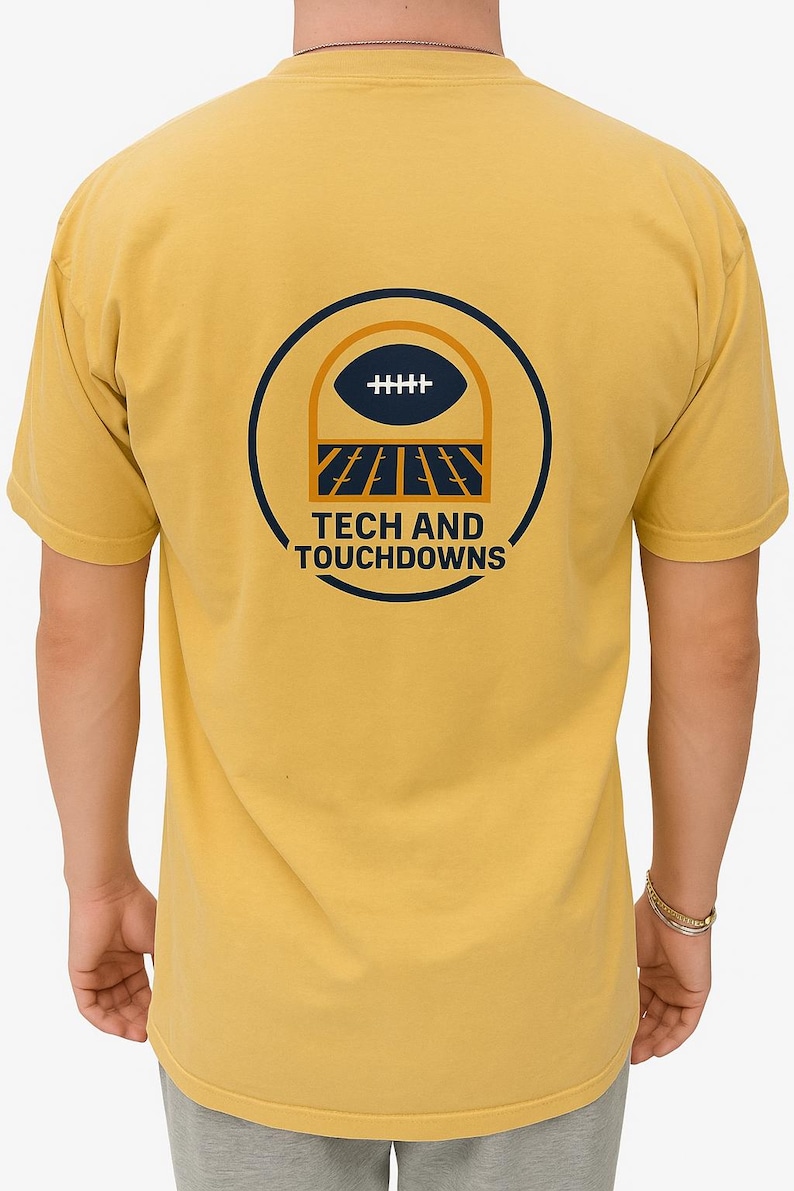 K&ouml;nnte beinhalten: Senfgelbes T-Shirt mit einem marineblauen und orangefarbenen Football-Motiv auf dem R&uuml;cken. Das Design zeigt einen Football, Torpfosten und den Text "TECH AND TOUCHDOWNS". Das T-Shirt hat kurze &Auml;rmel.