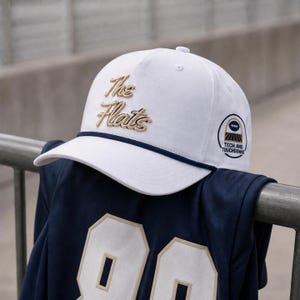 May include: A white baseball cap with a navy blue rope trim along the brim. The front features the text The Flats in gold embroidery. A circular patch on the side displays a football icon and the text Tech and Touchdowns in navy and gold.