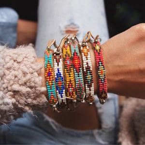 Puede incluir: Un conjunto de pulseras de cuentas coloridas con patrones geométricos. Las pulseras están hechas con cuentas pequeñas y coloridas y están enhebradas en un cordón de cuero. Las pulseras están apiladas en la muñeca de una persona.