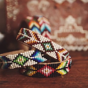 May include: Three beaded bracelets with colorful patterns on a brown wooden surface. The bracelets are made with small, colorful beads and have a leather base.