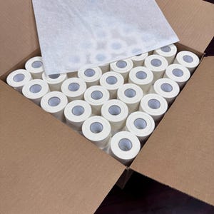 May include: A cardboard box filled with numerous rolls of white paper. The rolls are neatly arranged, with their circular ends visible, and are partially covered by a sheet of thin, white paper. The box is open, revealing the contents.