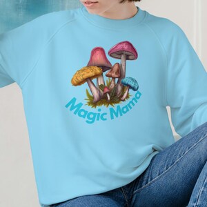 Könnte beinhalten: Hellblaues Sweatshirt mit einem farbenfrohen Pilz-Motiv. Das Design zeigt Pilze in Rot, Gelb, Braun und Blau, zusammen mit dem Text "Magic Mama". Ein bequemes und stilvolles Oberteil.
