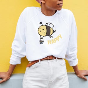 Könnte beinhalten: Ein weißes Langarm-Sweatshirt mit einer Cartoon-Bienen-Grafik, die einen kleinen grauen Eimer hält. Die Biene hat gelbe und schwarze Streifen und ein lächelndes Gesicht. Das Wort HAPPY ist in gelben Großbuchstaben neben der Biene gedruckt.