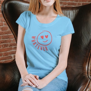 Könnte beinhalten: Ein hellblaues kurzärmeliges T-Shirt mit einer roten Grafik eines kreisförmigen Smileys mit herzförmigen Augen und dem Wort WHATEVER, das in roten Großbuchstaben unter dem Gesicht auf die Brust des Kleidungsstücks gedruckt ist.