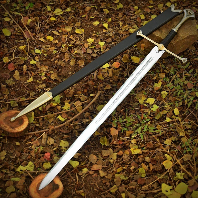 May include: A silver sword with a gold hilt and a black handle, resting on a bed of leaves. The sword has a long, sharp blade and is accompanied by a black sheath with gold accents. The sword is approximately 36 inches long.