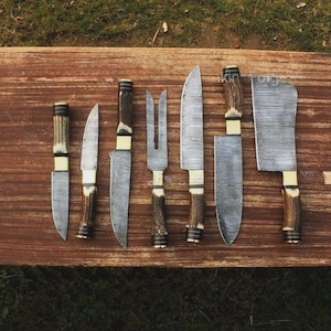 May include: A collection of handcrafted knives and a carving fork with textured blades and antler handles. The blades have a unique pattern, and the handles are accented with brass. The knives are arranged on a wooden surface.