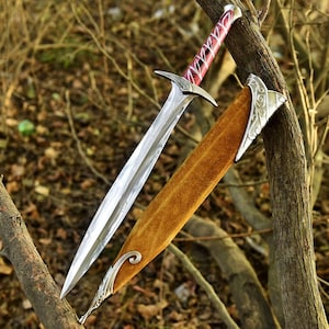 May include: A detailed image of a decorative sword with a silver blade and a red and silver handle. The sword is sheathed in a brown scabbard with silver accents. The sword is resting on a tree branch.