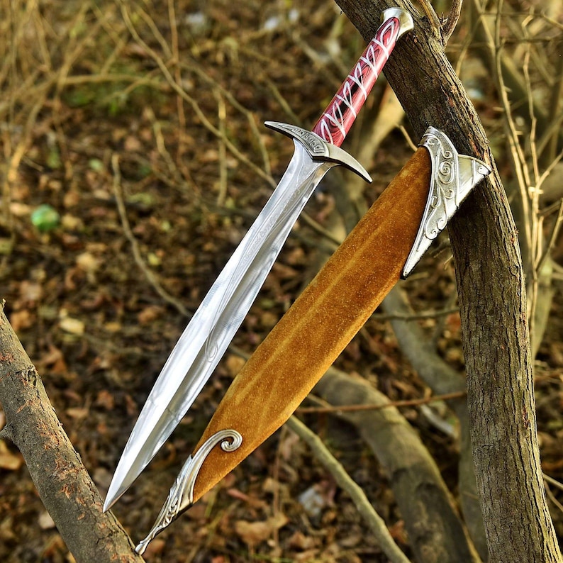 May include: A detailed image of a decorative sword with a silver blade and a red and silver handle. The sword is sheathed in a brown scabbard with silver accents. The sword is resting on a tree branch.