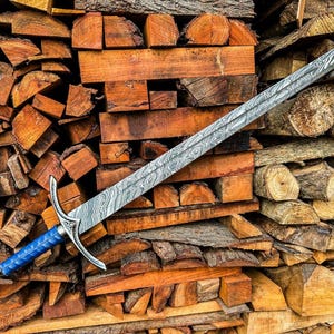 May include: A long, ornate sword with a patterned blade and a blue handle rests on a pile of firewood. The blade has a silver finish with a unique, swirling pattern. The handle is a vibrant blue, and the hilt is silver.