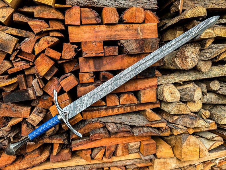 May include: A long, ornate sword with a patterned blade and a blue handle rests on a pile of firewood. The blade has a silver finish with a unique, swirling pattern. The handle is a vibrant blue, and the hilt is silver.