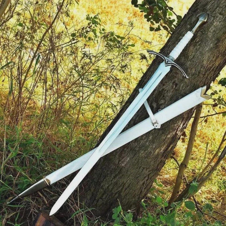 May include: A long, silver sword with a white handle and crossguard, resting in a white scabbard. The sword is leaning against a tree trunk, with a backdrop of green and yellow foliage. The blade is long and sharp, and the handle is wrapped in white material.