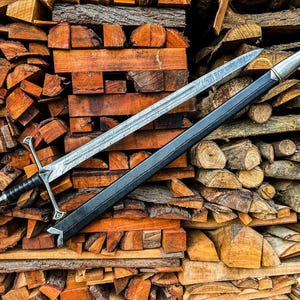 May include: A silver sword with a black handle and a black sheath rests on a pile of firewood. The sword has a detailed blade and ornate hilt. The wood is a mix of cut logs and split pieces, showing various shades of brown and orange.