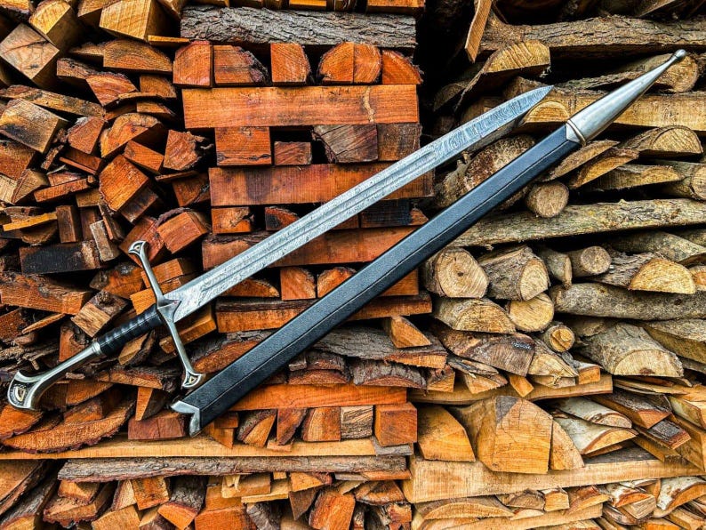 May include: A silver sword with a black handle and a black sheath rests on a pile of firewood. The sword has a detailed blade and ornate hilt. The wood is a mix of cut logs and split pieces, showing various shades of brown and orange.