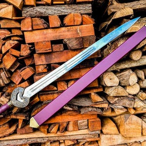 May include: A decorative sword with a silver blade and a purple handle and sheath. The handle has a decorative design. The sheath has gold-colored accents. The sword is displayed on a pile of wood.