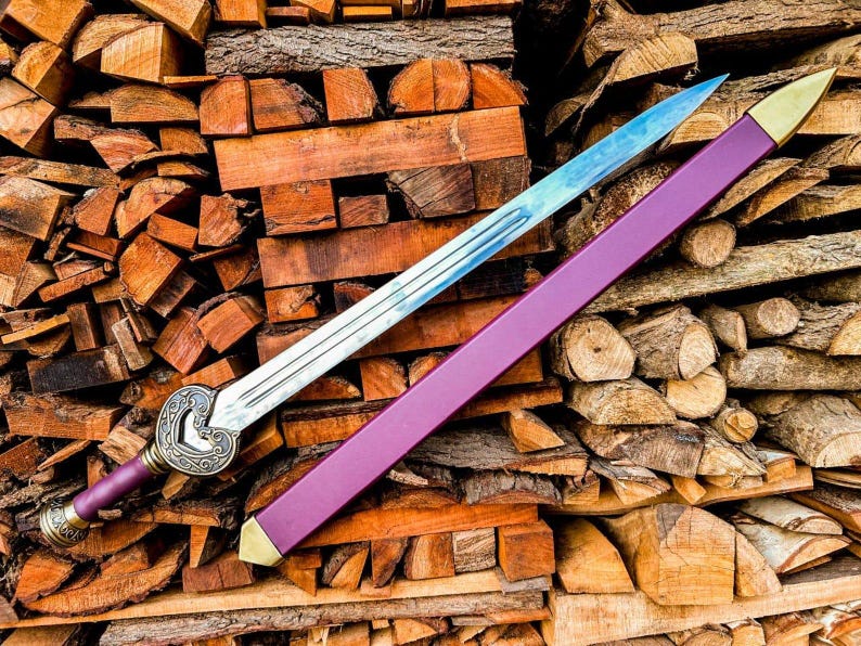 May include: A decorative sword with a silver blade and a purple handle and sheath. The handle has a decorative design. The sheath has gold-colored accents. The sword is displayed on a pile of wood.