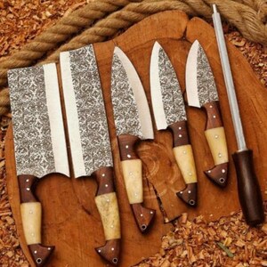 May include: A set of handcrafted kitchen knives with ornate patterned blades and wooden handles. The set includes a cleaver, several chef's knives, and a sharpening steel. The knives are displayed on a wooden cutting board.