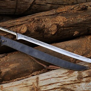 May include: A silver-colored sword with an ornate handle and a long, pointed blade rests on a bed of wooden logs. A black leather sheath with a brown strap accompanies the sword. The sword's blade features etched designs.