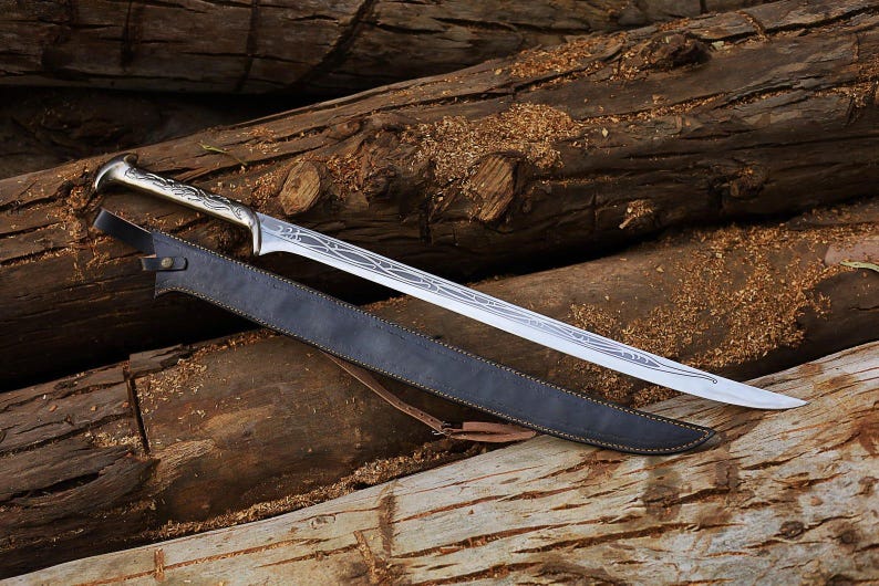 May include: A silver-colored sword with an ornate handle and a long, pointed blade rests on a bed of wooden logs. A black leather sheath with a brown strap accompanies the sword. The sword's blade features etched designs.