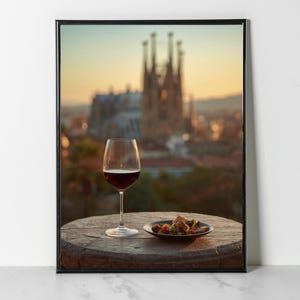 May include: A framed print featuring a wine glass filled with red wine and a small plate of food on a wooden table. The background shows a blurred cityscape with a large cathedral at sunset. The print is in a black frame.
