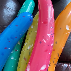 Könnte beinhalten: Nahaufnahme von mehreren langen, bunten Luftballons. Die Ballons sind in Blau-, Grün-, Gelb-, Rosa- und Orangetönen gehalten und mit weißen und lila Farbspritzern versehen. Die Ballons sind gebogen und scheinen aus einem glänzenden Material zu bestehen.