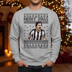 May include: Gray sweatshirt with a festive Christmas design. The graphic features a soccer player in a black and white striped jersey, with the text "MERRY XMAS" and "HO HO HOWAY THE LADS!" in white.