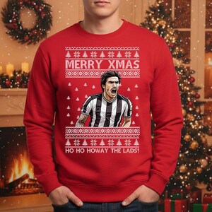 May include: Red Christmas sweater with a soccer player graphic. The sweater has the text "MERRY XMAS" and "HO HO HOWAY THE LADS!" in white. The soccer player is wearing a black and white striped jersey.
