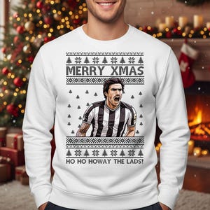 May include: White sweatshirt with a festive design. The sweatshirt features the text "MERRY XMAS" and "HO HO HOWAY THE LADS!" along with a graphic of a soccer player. The design includes snowflakes and Christmas trees.
