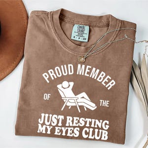 Op de afbeelding: Een bruin Comfort Colors T-shirt met de tekst "PROUD MEMBER OF THE JUST RESTING MY EYES CLUB" in het wit. Een zilveren ketting met een hanger en een bruine hoed zijn ook zichtbaar.
