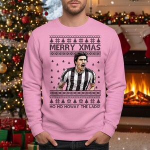 May include: A light pink Christmas sweater with the text "MERRY XMAS" and "HO HO HOWAY THE LADS!". The sweater features a graphic of a soccer player in a black and white striped jersey. The sweater has a festive design with Christmas trees.