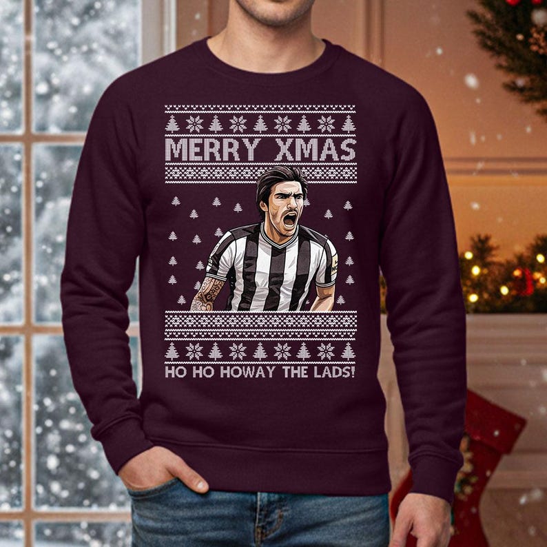 May include: A burgundy sweatshirt with a festive design. The text "MERRY XMAS" is above a graphic of a soccer player in a black and white striped jersey. Below the graphic is the text "HO HO HOWAY THE LADS!" White Christmas trees and snowflakes are also featured.