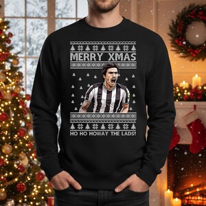 May include: Black sweatshirt with a festive design. The text "MERRY XMAS" is above a graphic of a soccer player in a black and white striped jersey. The text "HO HO HOWAY THE LADS!" is below the graphic.