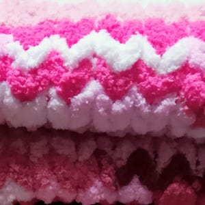 Puede incluir: Una manta plegada en tonos rosa, blanco y rosa. La manta presenta un patrón en zigzag texturizado en diferentes tonos de rosa y blanco, creando una apariencia suave y acogedora. La manta está hecha de un material grueso y esponjoso.